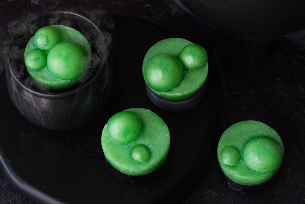 Melt and pour soap bars shaped like round bubbling cauldrons. The bottom layer is dyed black and the top layer is made up of green soap spheres to look like bubbles.